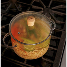 Clear glass cooking pot with metal lid and bamboo knob, filled with spaghetti and vegetables.
