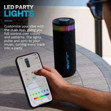 Black portable speaker with RGB top ring and blue logo, held near a smartphone.