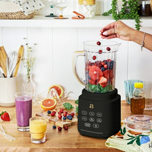 Black blender with clear pitcher holding berries and greens, next to cups of smoothie