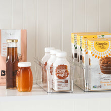 Clear plastic pantry bin with front handle opening, holding bottles and packaged foods.