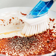 Dishwashing brush with blue handle scrubbing red food stains in a pan