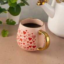 Pink ceramic mug with red heart pattern and gold-tone handle, dark drink inside.
