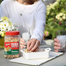 Jar of oven-roasted cashews on table with bowl and hand adding cashews