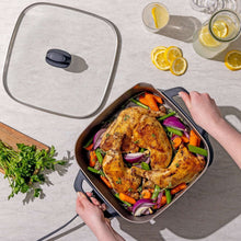 Ceramic electric skillet with glass lid open, hands placing seasoned chicken and vegetables