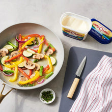 Spreadable butter tub with spoon, beside cooked vegetables and a chef’s knife