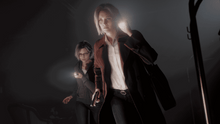 PS5 game action scene with two women in dark clothing, one reaching for a light.