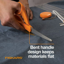 Orange-handled Fiskars scissors cutting fabric on a surface, showing long steel blades.