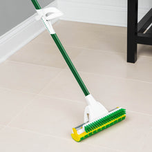 Sponge mop with scrub brush and green handle, yellow bristle edge on tiled floor.