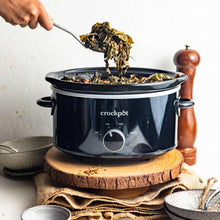 Black slow cooker with lid open, spoon lifting food, and front dial visible