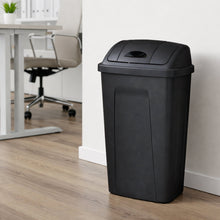 Black plastic trash can with swing-top lid, upright rectangular shape on floor.