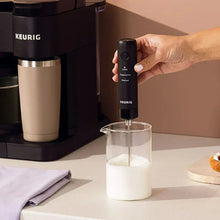 Black milk frother held by hand with whisk submerged in a cup of milk