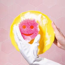 Yellow cleaning sponge with pink scrub face held by a white-gloved hand