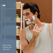 Man shaving with white shave cream on face, holding razor; light gray sleeveless shirt