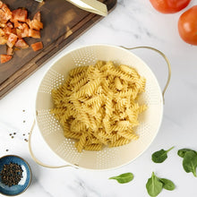 Cream perforated colander holding dry pasta, with gold rim handles.