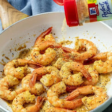 Seasoned shrimp in a skillet with lemon pepper seasoning sprinkled from a red-capped shaker