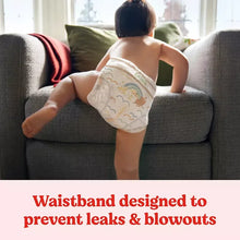 Baby standing wearing white printed diapers on a gray couch, seen from behind.