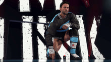 Wrestler kneeling in arena wearing dark jacket with blue armbands