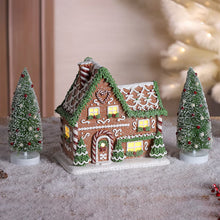 Prelit gingerbread house decoration with two snowy pine trees, warm lit windows