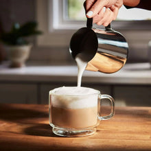 Glass mug of foamy coffee with crema being poured from a metal jug