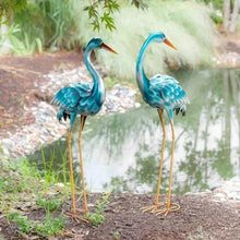 Two teal heron metal statues with orange beaks and gold wire legs, standing close.