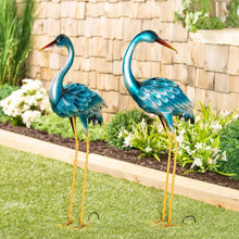 Two teal heron metal sculptures with orange beaks and gold wire legs, posed upright.