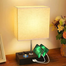 Lit fabric-shade bedside lamp with black charging base and phone on a stand
