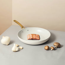 White ribbed fry pan with gold handle with steak, garlic, and mushrooms