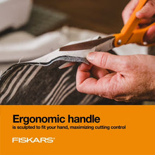 Fiskars orange-handled scissors in use, cutting dark fabric with silver blades.