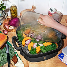 Electric skillet with glass lid, holding vegetables and herbs in a black pan