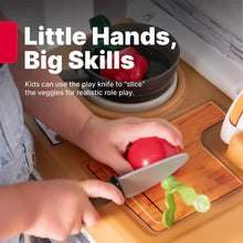 Hands slicing pretend red pepper on wooden cutting board beside toy stove