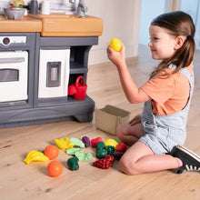 Toy play kitchen set with stove and utensils; child holds a yellow lemon prop