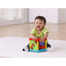Busy learners activity cube toy in front of baby; red top, yellow frame, blue base