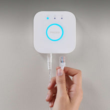Wall-mounted Philips Hue bridge hub with blue ring as a hand plugs in Ethernet cable