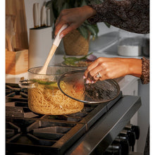 Clear glass pot on stovetop with side handles, bamboo-handled utensil over contents.