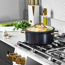 Black non-stick stock pot with glass lid and gold-tone handles, simmering food
