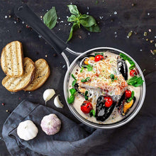 Stainless saucepan with black handles, filled with seafood and herbs