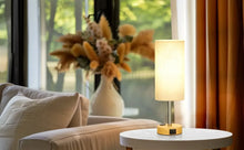 Small bedside table lamp with fabric drum shade and round wood base, lit glow