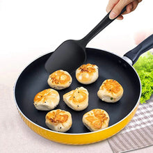 Black silicone spatula with wide rounded head, used in a skillet to lift food.