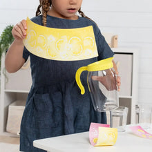 Child holding a lemonade pretend play toy pitcher above cups on a table