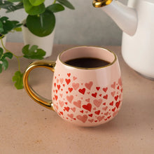 Pink ceramic mug with red heart pattern and gold-tone handle, dark drink inside.