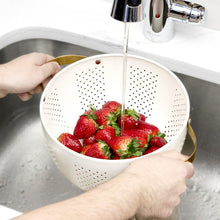 Cream colander with gold handles, filled with strawberries under running water.