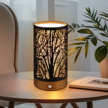 Table lamp with black cylindrical branch-pattern shade and wooden base on a bedside table.