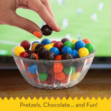 Clear bowl of multicolor candy-coated chocolates with pretzel pieces, hand adding candies