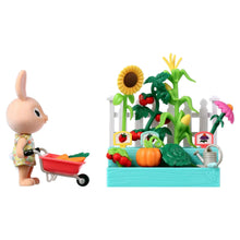 Bunny figure holding wheelbarrow with carrot and leafy greens beside raised garden box