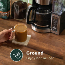 Glass mug of ground coffee in hand next to a coffee maker, hot or iced