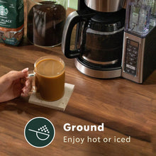 Hand holding a glass mug of coffee next to a coffee maker