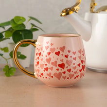 Pink ceramic mug with red heart pattern and gold-tone handle, dark interior.
