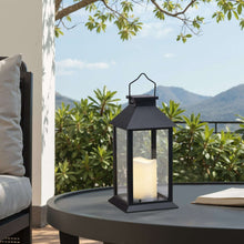 Black solar lantern with lit candle inside, glass panels and metal handle on patio.