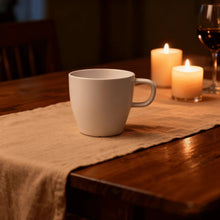 White coffee mug with curved handle on a cloth-covered wooden table.