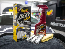 Car clay kit box, red spray bottle, clay bars, and yellow microfiber cloth on car surface